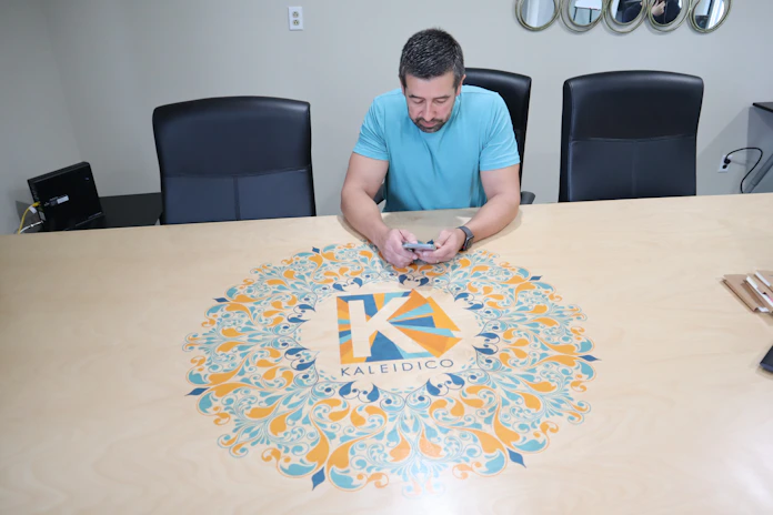 A man in a light blue shirt is seated at a large conference table, which has a colorful circular design featuring an intricate pattern and the word 'Kaleidico' in the center. The table is set in a modern office environment with black chairs, a wall with mirrors, and electronic equipment in the background.