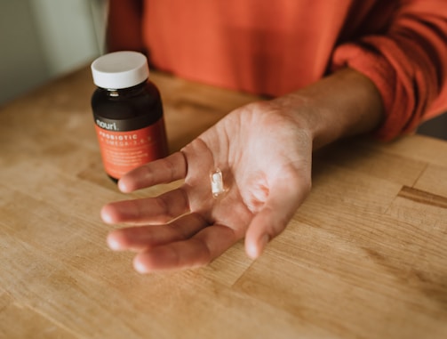 A hand is extended over a wooden surface, holding a single capsule. To the left, there's a dark bottle with a red label, which appears to be a supplement or vitamin container.