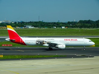 A commercial airplane featuring Iberia livery with additional promotional graphics is on the tarmac at an airport. The scene includes a green grassy area surrounding the runway, and in the distant background, trees and urban structures are visible under a clear blue sky.