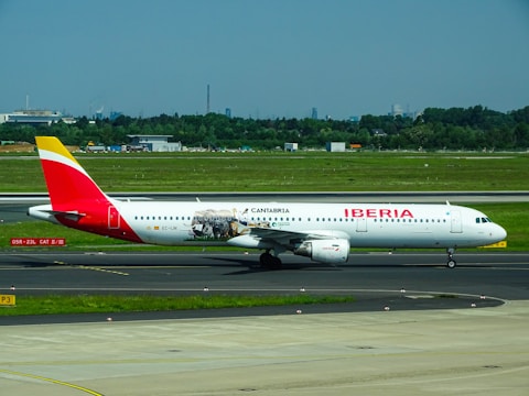 A commercial airplane featuring Iberia livery with additional promotional graphics is on the tarmac at an airport. The scene includes a green grassy area surrounding the runway, and in the distant background, trees and urban structures are visible under a clear blue sky.