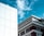 The image features two buildings contrasted against a clear blue sky with scattered clouds. The left side displays a modern white building with a grid of clean lines and geometric panels. On the right, a more traditional building showcases ornate architectural details with decorative moldings and a red brick facade.