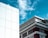 The image features two buildings contrasted against a clear blue sky with scattered clouds. The left side displays a modern white building with a grid of clean lines and geometric panels. On the right, a more traditional building showcases ornate architectural details with decorative moldings and a red brick facade.