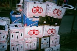Stacks of cardboard boxes are piled on a sidewalk. The boxes have various labels and logos, some featuring red designs with symbols and others with blue. There are some visible brands like Bally and Patron, and the boxes appear to contain produce. In the background, a parked car and other urban features can be seen.