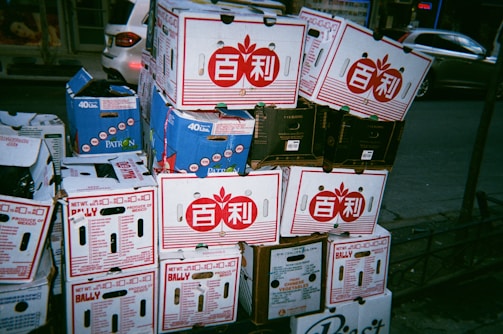 Stacks of cardboard boxes are piled on a sidewalk. The boxes have various labels and logos, some featuring red designs with symbols and others with blue. There are some visible brands like Bally and Patron, and the boxes appear to contain produce. In the background, a parked car and other urban features can be seen.