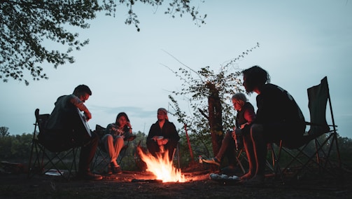 A group of campers sitting around a campfire at dusk in a forest clearing.