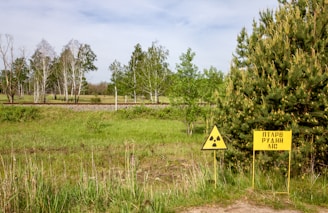A grassy field with sparse trees, some of which have bare branches, is shown under a partly cloudy sky. There are two yellow warning signs on the right side of the field; one of them features a radiation symbol and the other has text. In the background, there's a metal pole and a set of train tracks running horizontally through the scene.