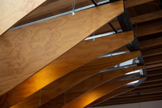 A close-up of wooden slats or panels forming a ceiling structure. The wooden elements have a natural finish with visible wood grain patterns and are supported by metallic braces. The panels are partly illuminated by a warm light source, creating a cozy atmosphere.