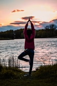 A person practices a yoga pose, standing on one leg with hands raised above the head, near the edge of a body of water during sunset. The background includes trees and the sky filled with soft hues of orange, yellow, and pink clouds.