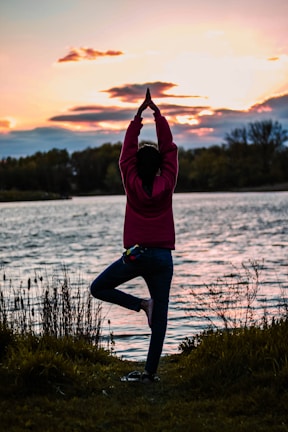 A person practices a yoga pose, standing on one leg with hands raised above the head, near the edge of a body of water during sunset. The background includes trees and the sky filled with soft hues of orange, yellow, and pink clouds.