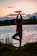 A person practices a yoga pose, standing on one leg with hands raised above the head, near the edge of a body of water during sunset. The background includes trees and the sky filled with soft hues of orange, yellow, and pink clouds.