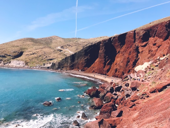 A coastal landscape featuring a beach with vibrant turquoise waters meeting rugged red cliffs. The steep cliffs have a rich, earthy hue, contrasting with the clear blue sky above. Waves gently crash against the shoreline, and a few people are visible on the beach, adding a sense of scale to the dramatic scenery.