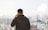 A person stands with their back turned, talking on a mobile phone. They overlook a city skyline through a large window, with tall buildings visible in the distance. The atmosphere appears calm and contemplative.