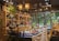 A well-lit interior of a shop filled with a variety of pottery and ceramic items, displayed on shelves and tables. The shop showcases an array of teapots, bowls, and decorative statues. The ambiance is enhanced by hanging lamps and a wooden slatted ceiling. There's a touch of greenery with potted plants adding to the decor, and large windows offer a view of trees outside.