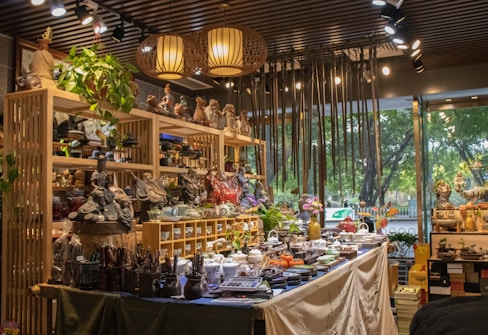 A well-lit interior of a shop filled with a variety of pottery and ceramic items, displayed on shelves and tables. The shop showcases an array of teapots, bowls, and decorative statues. The ambiance is enhanced by hanging lamps and a wooden slatted ceiling. There's a touch of greenery with potted plants adding to the decor, and large windows offer a view of trees outside.