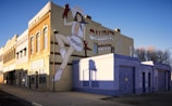 A large mural painted on the side of a building, depicting a person in western attire with a headdress and a high-kick pose. The mural is accompanied by text indicating a greeting from Calumet. The building facade features traditional architectural details and is bathed in warm sunlight. Shadows and a clear blue sky are visible, adding to the vibrant yet serene atmosphere.