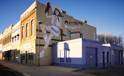 A large mural painted on the side of a building, depicting a person in western attire with a headdress and a high-kick pose. The mural is accompanied by text indicating a greeting from Calumet. The building facade features traditional architectural details and is bathed in warm sunlight. Shadows and a clear blue sky are visible, adding to the vibrant yet serene atmosphere.
