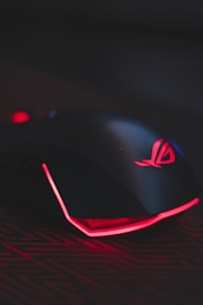 A sleek gaming mouse with a black finish and red backlighting. The mouse features a glowing logo on the top and an illuminated edge, placed on a surface with a subtle circuit-like pattern.