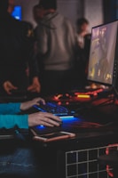A person is engaged in a video game at a computer setup, using a keyboard and mouse. The scene is dimly lit, with the glow from the monitor reflecting off the hands of the player. Several other people are present in the background, indicating a social or competitive gaming environment.