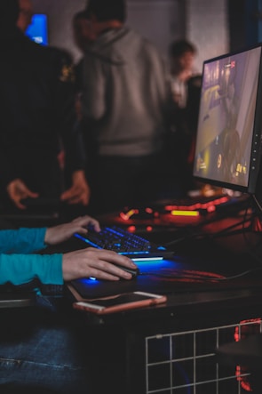 A person is engaged in a video game at a computer setup, using a keyboard and mouse. The scene is dimly lit, with the glow from the monitor reflecting off the hands of the player. Several other people are present in the background, indicating a social or competitive gaming environment.
