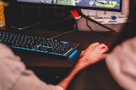 Two people are seated at a computer desk engaged in a video game displayed on a monitor. The setup includes a mechanical keyboard with glowing blue backlighting. The environment has a casual, gaming setup with a can of beverage in the background.