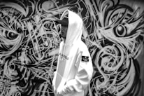 A person wearing a white hoodie and cap stands in front of a detailed black-and-white graffiti wall. The graffiti features intricate abstract designs with swirling patterns and eye-like shapes.