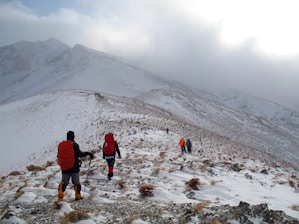 A group of hikers with backpacks walking along a snow-covered mountain trail. The landscape is vast with rolling hills in the background, partially obscured by clouds. The ground is rocky with patches of snow, and the sky is overcast, adding an element of challenge to the climb.