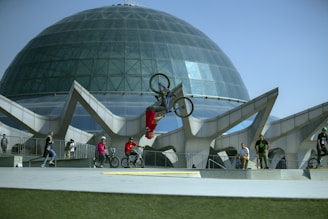 A BMX rider performs a backflip in front of a large, modern glass dome structure. Other bikers and onlookers, wearing casual clothing and helmets, watch the performance. The architecture features geometric designs and a clear blue sky is visible in the background.