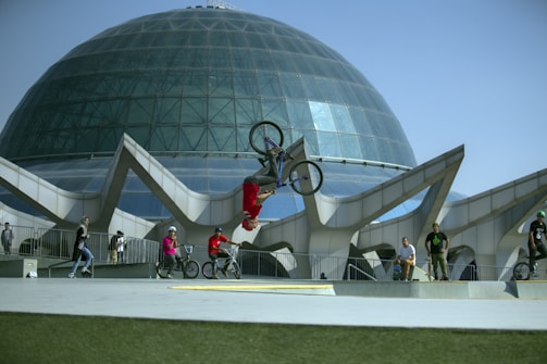 A BMX rider performs a backflip in front of a large, modern glass dome structure. Other bikers and onlookers, wearing casual clothing and helmets, watch the performance. The architecture features geometric designs and a clear blue sky is visible in the background.