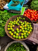 A vibrant market scene featuring a variety of fresh vegetables and fruits. A wicker basket is filled with green lemons, surrounded by baskets of bright red tomatoes, green spinach leaves, and long green beans. A turquoise box contains bitter gourds, and another sack is filled with green, cucumber-like vegetables.