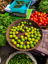 A vibrant market scene featuring a variety of fresh vegetables and fruits. A wicker basket is filled with green lemons, surrounded by baskets of bright red tomatoes, green spinach leaves, and long green beans. A turquoise box contains bitter gourds, and another sack is filled with green, cucumber-like vegetables.