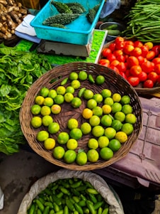 A vibrant market scene featuring a variety of fresh vegetables and fruits. A wicker basket is filled with green lemons, surrounded by baskets of bright red tomatoes, green spinach leaves, and long green beans. A turquoise box contains bitter gourds, and another sack is filled with green, cucumber-like vegetables.