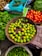 A vibrant market scene featuring a variety of fresh vegetables and fruits. A wicker basket is filled with green lemons, surrounded by baskets of bright red tomatoes, green spinach leaves, and long green beans. A turquoise box contains bitter gourds, and another sack is filled with green, cucumber-like vegetables.