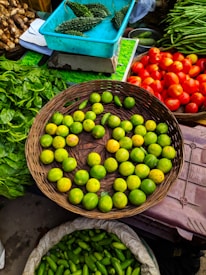 A vibrant market scene featuring a variety of fresh vegetables and fruits. A wicker basket is filled with green lemons, surrounded by baskets of bright red tomatoes, green spinach leaves, and long green beans. A turquoise box contains bitter gourds, and another sack is filled with green, cucumber-like vegetables.