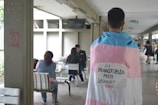 A person is standing indoors wearing a flag with the colors pink, white, and blue draped over their shoulders. There is a sign attached to the flag with the words 'La TRANSFOBIA MATA ¡CÚRATE!' taped on it. In the background, a few people are sitting at tables, suggesting a casual or communal area.
