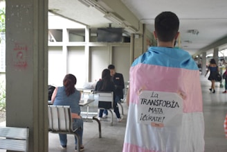 A person is standing indoors wearing a flag with the colors pink, white, and blue draped over their shoulders. There is a sign attached to the flag with the words 'La TRANSFOBIA MATA ¡CÚRATE!' taped on it. In the background, a few people are sitting at tables, suggesting a casual or communal area.