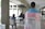 A person is standing indoors wearing a flag with the colors pink, white, and blue draped over their shoulders. There is a sign attached to the flag with the words 'La TRANSFOBIA MATA ¡CÚRATE!' taped on it. In the background, a few people are sitting at tables, suggesting a casual or communal area.