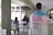 A person is standing indoors wearing a flag with the colors pink, white, and blue draped over their shoulders. There is a sign attached to the flag with the words 'La TRANSFOBIA MATA ¡CÚRATE!' taped on it. In the background, a few people are sitting at tables, suggesting a casual or communal area.