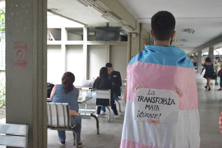 A person is standing indoors wearing a flag with the colors pink, white, and blue draped over their shoulders. There is a sign attached to the flag with the words 'La TRANSFOBIA MATA ¡CÚRATE!' taped on it. In the background, a few people are sitting at tables, suggesting a casual or communal area.