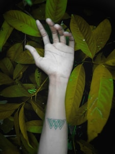 A human hand with a geometric tattoo on the wrist is extended in front of a background of large green leaves. The lighting casts a soft shadow, creating a contrast between the hand and the foliage.