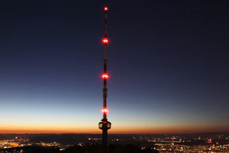 A sleek black and red telecommunications tower glowing at dusk against a city skyline.
