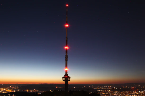 A sleek black and red telecommunications tower glowing at dusk against a city skyline.
