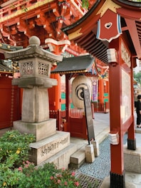 An ornate Japanese shrine setting featuring traditional red wooden structures with intricate architectural details. A stone lantern stands prominently beside lush green plants. Decorative elements include a large paper lantern and ornate carvings. The scene is vibrant and culturally rich.