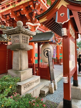 An ornate Japanese shrine setting featuring traditional red wooden structures with intricate architectural details. A stone lantern stands prominently beside lush green plants. Decorative elements include a large paper lantern and ornate carvings. The scene is vibrant and culturally rich.