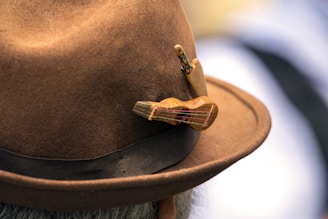 A close-up of a felt hat adorned with a small wooden miniature guitar and a wooden stick figure. The hat is brown with a dark band around the base.