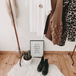 A cozy corner featuring a clothing rack with various garments, including a white t-shirt with an emblem and a range of sweaters. Below the rack on a white fluffy rug are a pair of black boots, a potted cactus, and a framed motivational quote that reads 'Get up, dress up, show up, never give up'. To the left, there is a tall decorative plant in a clear vase.