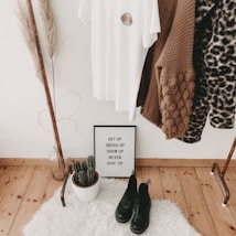 A cozy corner featuring a clothing rack with various garments, including a white t-shirt with an emblem and a range of sweaters. Below the rack on a white fluffy rug are a pair of black boots, a potted cactus, and a framed motivational quote that reads 'Get up, dress up, show up, never give up'. To the left, there is a tall decorative plant in a clear vase.