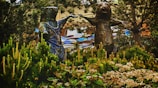 Two anthropomorphic animal sculptures, possibly bears, are positioned among lush greenery, appearing to be holding hands. The scene includes a variety of plants and trees surrounding the statues, and a rhythmic, patterned background with a mix of natural and artistic elements.
