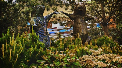 Two anthropomorphic animal sculptures, possibly bears, are positioned among lush greenery, appearing to be holding hands. The scene includes a variety of plants and trees surrounding the statues, and a rhythmic, patterned background with a mix of natural and artistic elements.