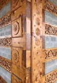 Intricate wooden carvings adorn a corner of a structure, featuring circular patterns and floral designs. The carvings are set against a backdrop of horizontally arranged stone tiles with a textured surface.