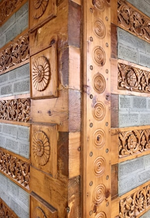 Intricate wooden carvings adorn a corner of a structure, featuring circular patterns and floral designs. The carvings are set against a backdrop of horizontally arranged stone tiles with a textured surface.